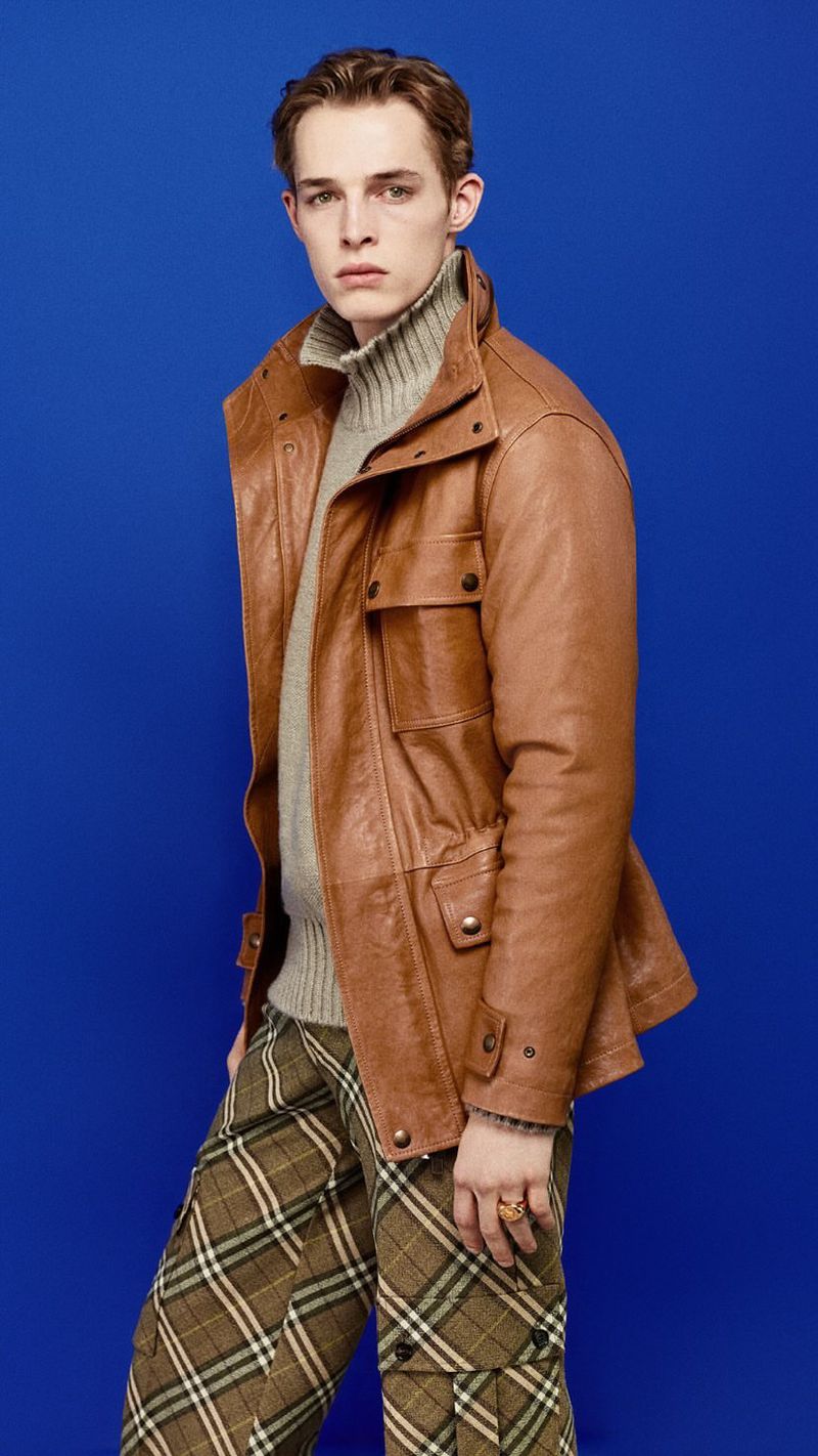 A young man poses indoors wearing a tan leather jacket, grey roll-neck sweater, and plaid pants against a blue background.