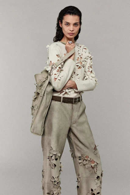 Brunette woman in light green floral-embellished sweater and pants holding matching tote bag on a neutral backdrop in a studio shot.