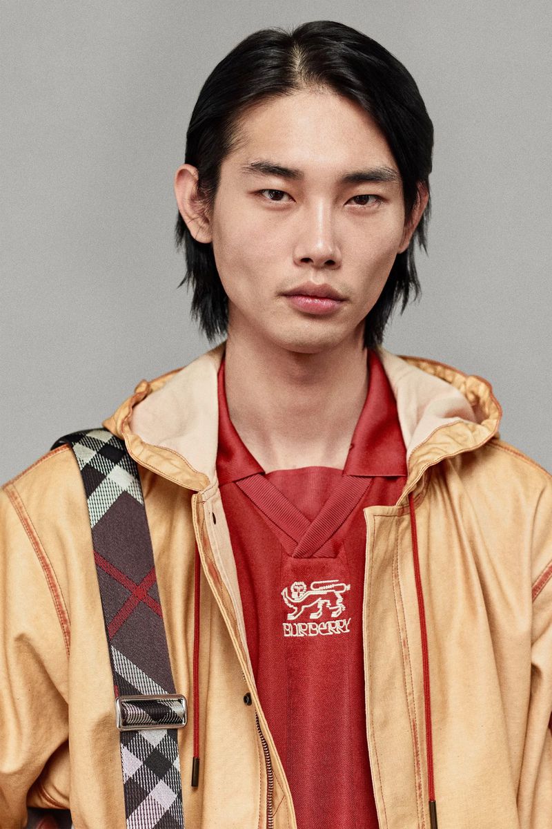 Close-up of an Asian male fashion model with black hair wearing a Burberry plaid strap, jacket and top against a grey background.
