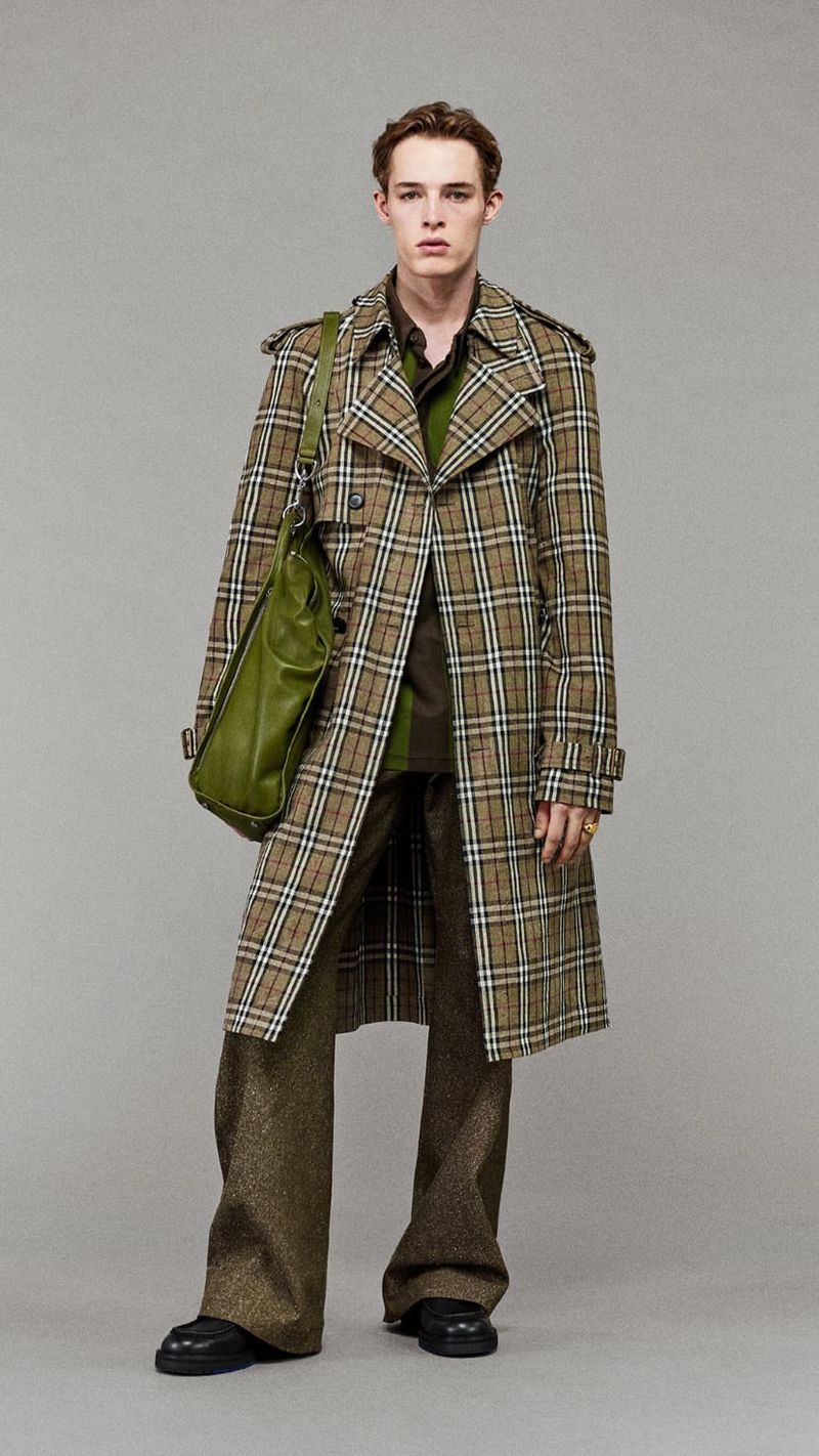 Full shot of a young adult male fashion model posing in a brown plaid trench coat with an olive green shoulder bag in a photo studio.