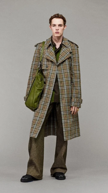 Full shot of a young adult male fashion model posing in a brown plaid trench coat with an olive green shoulder bag in a photo studio.