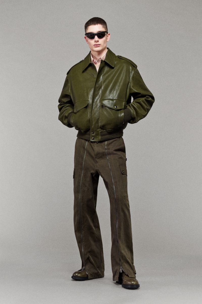 Male fashion model poses in green bomber jacket and olive pants against a neutral backdrop, showcasing modern military style.