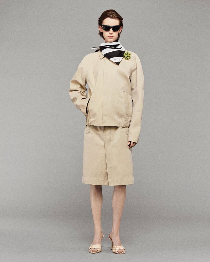 Woman in beige jacket and skirt, black and white scarf, sunglasses, and heels, posing in a full shot against a grey studio backdrop.