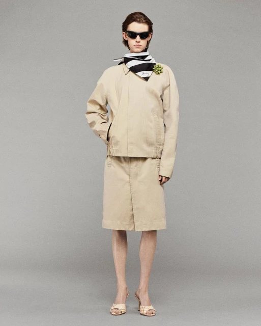 Woman in beige jacket and skirt, black and white scarf, sunglasses, and heels, posing in a full shot against a grey studio backdrop.