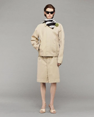 Woman in beige jacket and skirt, black and white scarf, sunglasses, and heels, posing in a full shot against a grey studio backdrop.