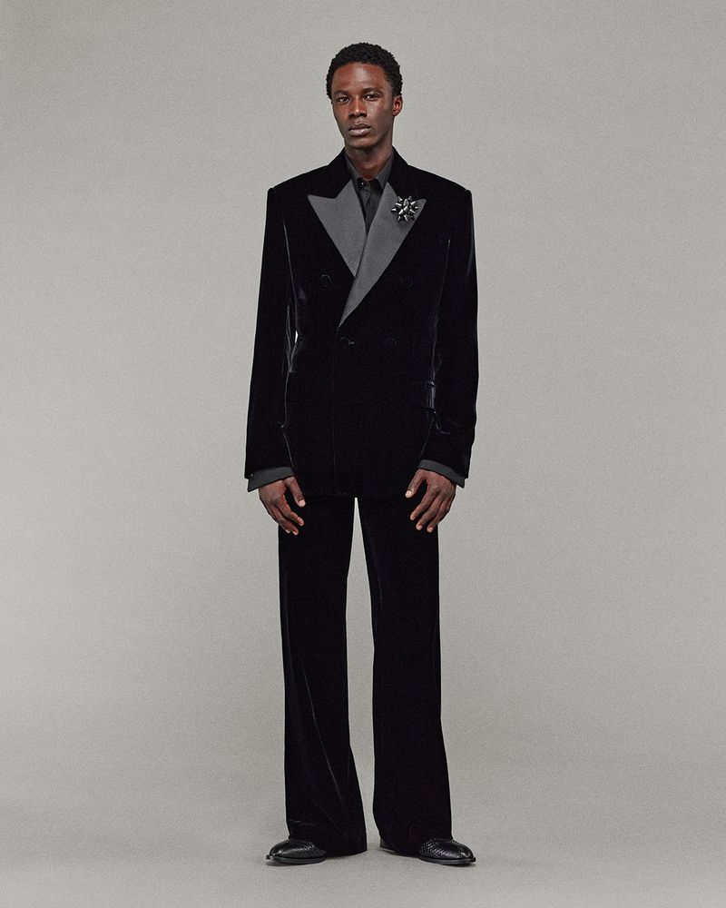 An adult male model stands in a black velvet double-breasted suit with wide-legged pants, accessorized with a lapel pin on a grey background.