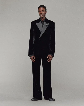 An adult male model stands in a black velvet double-breasted suit with wide-legged pants, accessorized with a lapel pin on a grey background.