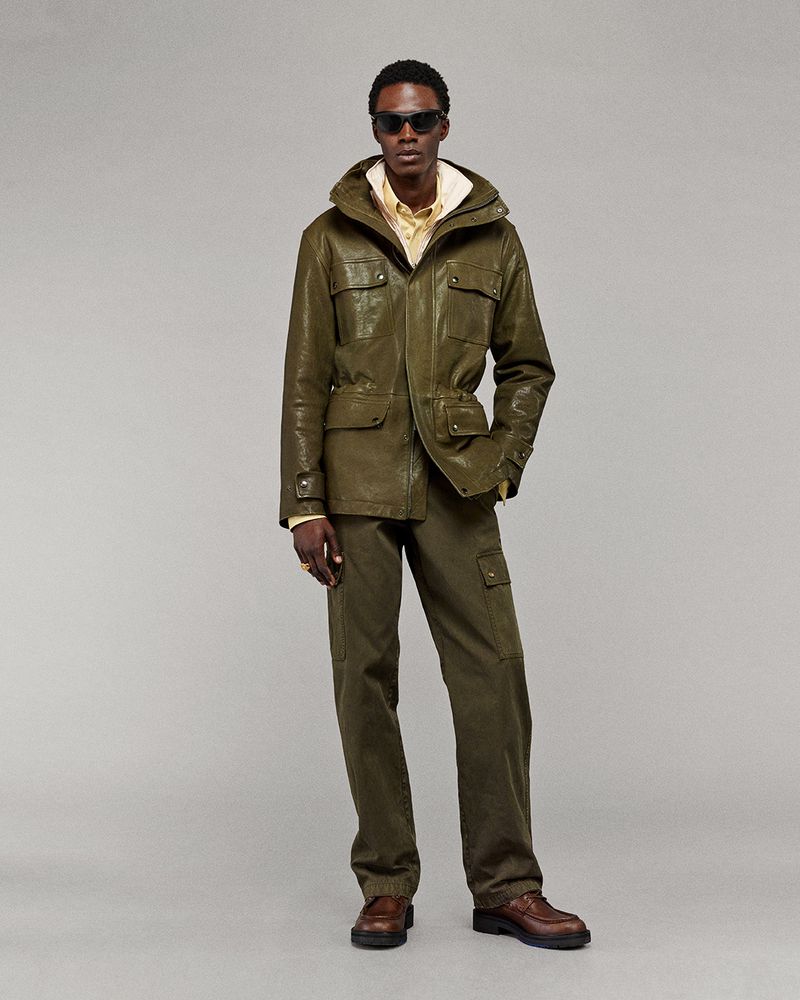 A full-shot studio portrait of a Black male model showcasing an olive-green hooded jacket and pants with brown leather dress shoes on a light backdrop.