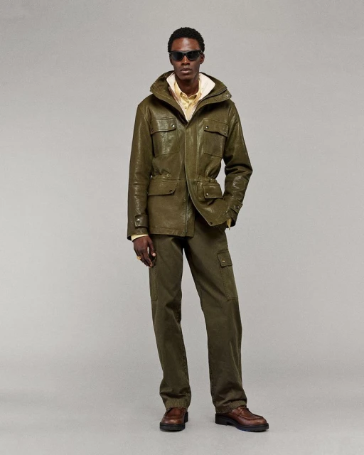 A full-shot studio portrait of a Black male model showcasing an olive-green hooded jacket and pants with brown leather dress shoes on a light backdrop.