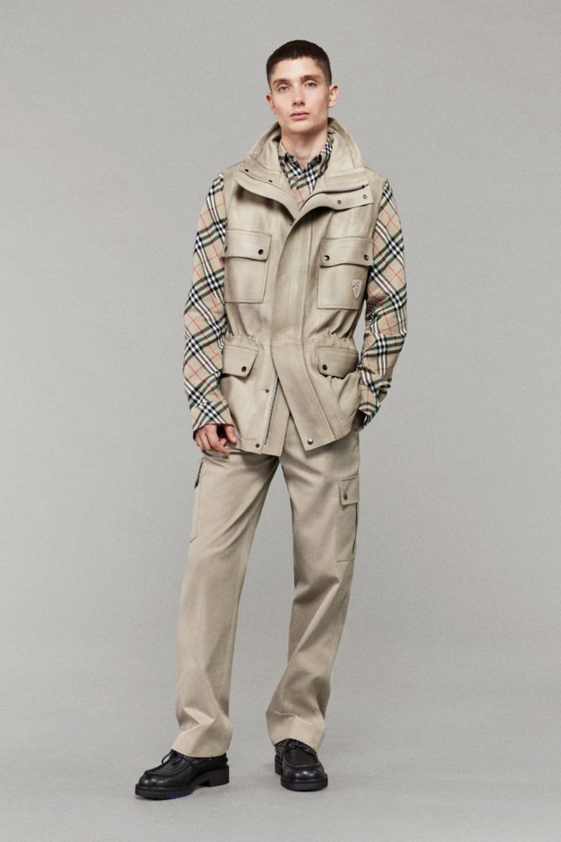 Full-length studio shot of a male model wearing a beige vest, patterned shirt, and cargo pants. He is posing against a grey background.