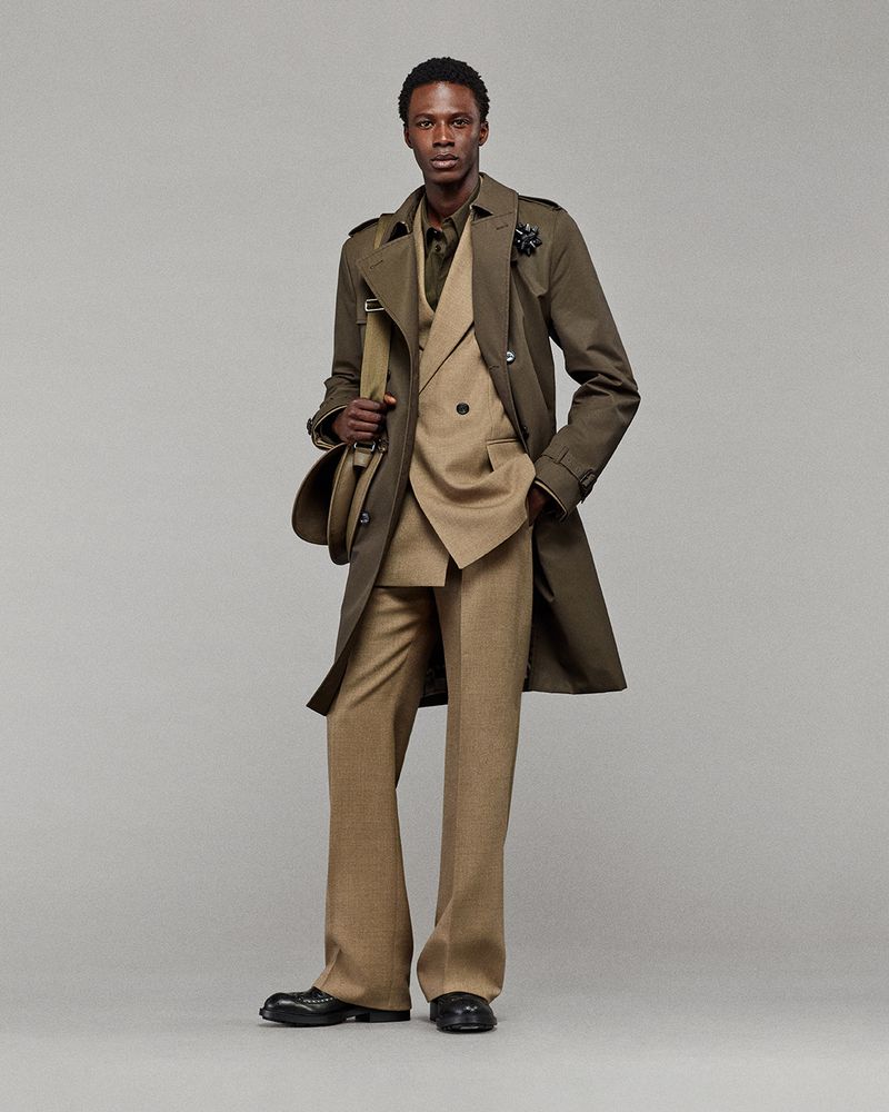 Full-length shot of a Black man in a double-breasted suit with a trench coat and shoulder bag, posing in a neutral studio setting. Fashion.