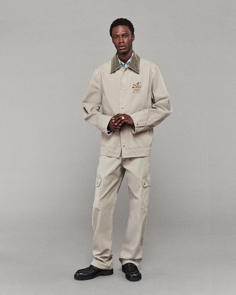 Man posing in a beige workwear jacket and cargo pants against a grey background, showcasing fashion-forward style and contemporary menswear.