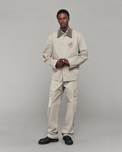 Man posing in a beige workwear jacket and cargo pants against a grey background, showcasing fashion-forward style and contemporary menswear.