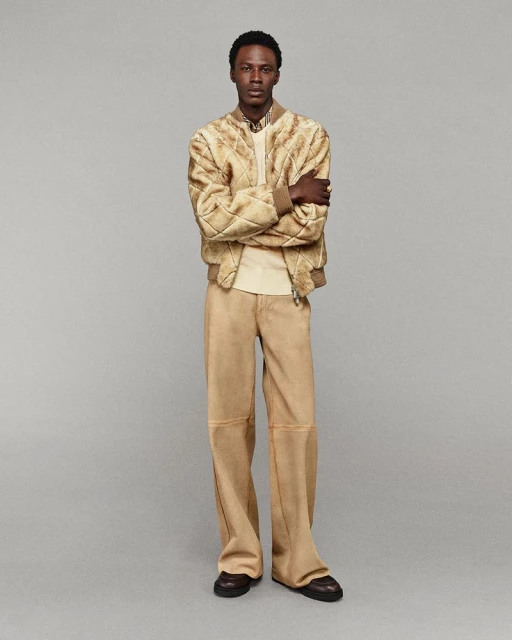 A full shot of a Black male fashion model in a tan bomber jacket and wide-leg pants, posing against a neutral gray studio background.