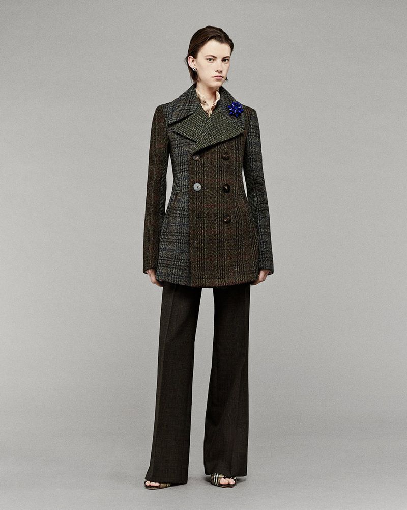A fashionably dressed woman in a mixed-fabric pea coat poses in a studio setting with a neutral background, wearing wide-leg trousers and sandals.
