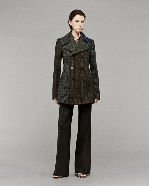 A fashionably dressed woman in a mixed-fabric pea coat poses in a studio setting with a neutral background, wearing wide-leg trousers and sandals.
