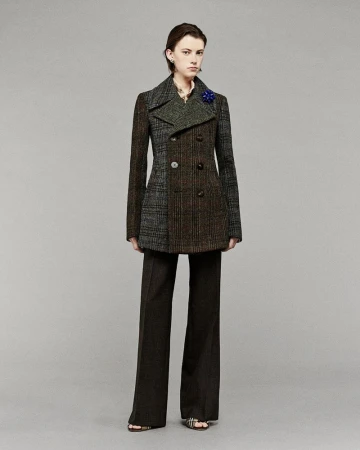 A fashionably dressed woman in a mixed-fabric pea coat poses in a studio setting with a neutral background, wearing wide-leg trousers and sandals.