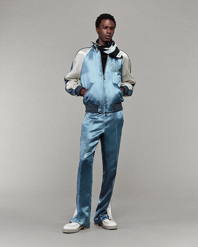 Dark-skinned male fashion model poses in light blue satin jacket and pants, with a black and white scarf, in a neutral studio.