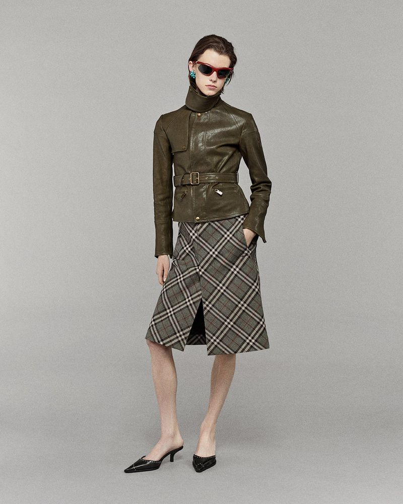 A stylish adult female poses in a photo studio, wearing a belted leather jacket, plaid skirt, and black heeled shoes against a grey backdrop.