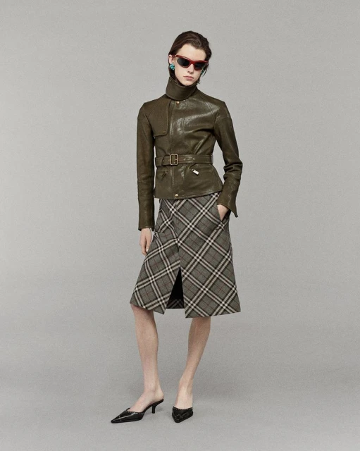 A stylish adult female poses in a photo studio, wearing a belted leather jacket, plaid skirt, and black heeled shoes against a grey backdrop.