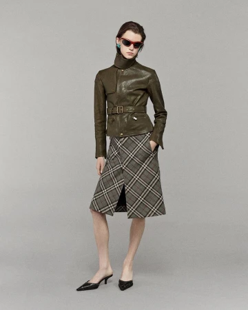 A stylish adult female poses in a photo studio, wearing a belted leather jacket, plaid skirt, and black heeled shoes against a grey backdrop.