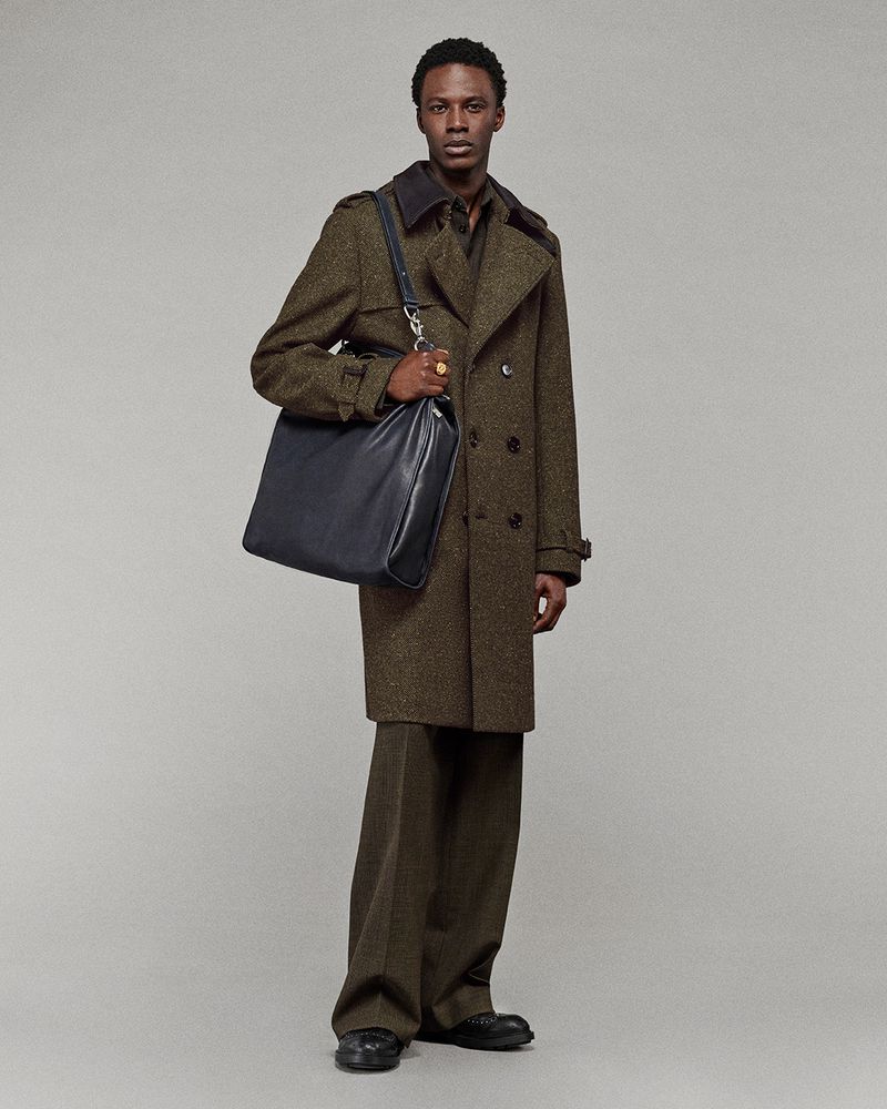 Man in herringbone overcoat holding blue leather bag in full-length studio shot with muted gray background, fashion and style editorial.