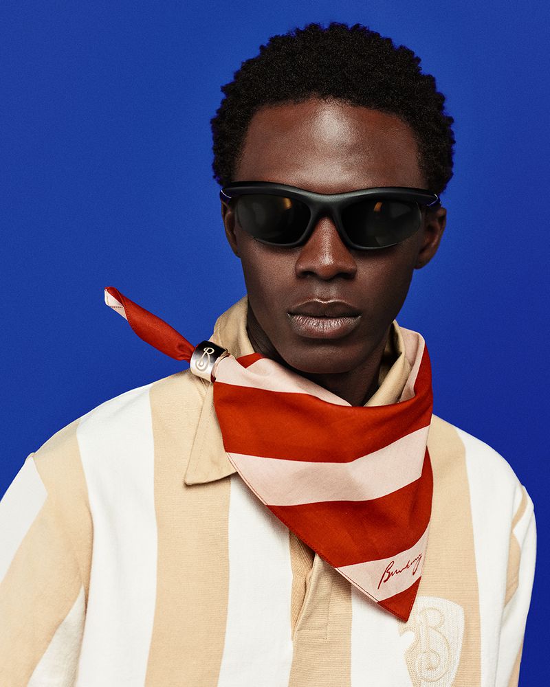 Portrait of a dark-skinned male model wearing sunglasses and a striped shirt, accessorized with a red and white striped scarf, on blue background.