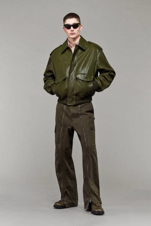 Male model wearing an olive green leather bomber jacket, cargo trousers with front zippers, and sunglasses against a grey backdrop.