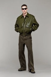 Male model wearing an olive green leather bomber jacket, cargo trousers with front zippers, and sunglasses against a grey backdrop.