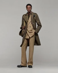 A Black male model stands wearing a beige double-breasted suit beneath an olive green trench coat against a solid grey studio backdrop.
