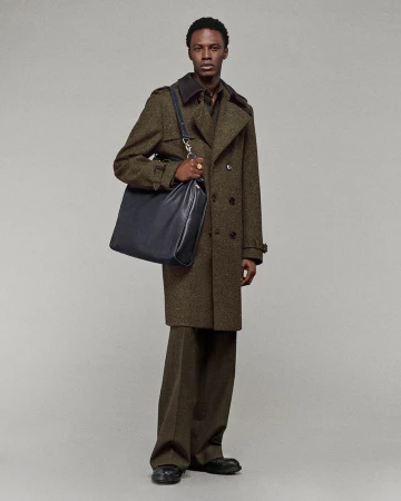 Model Cherif Douamba wearing an olive green trench coat and carrying a dark leather shoulder bag against a grey studio backdrop.