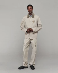 Model wearing a beige collared utility jacket and matching cargo trousers with black dress shoes against a solid grey studio background.