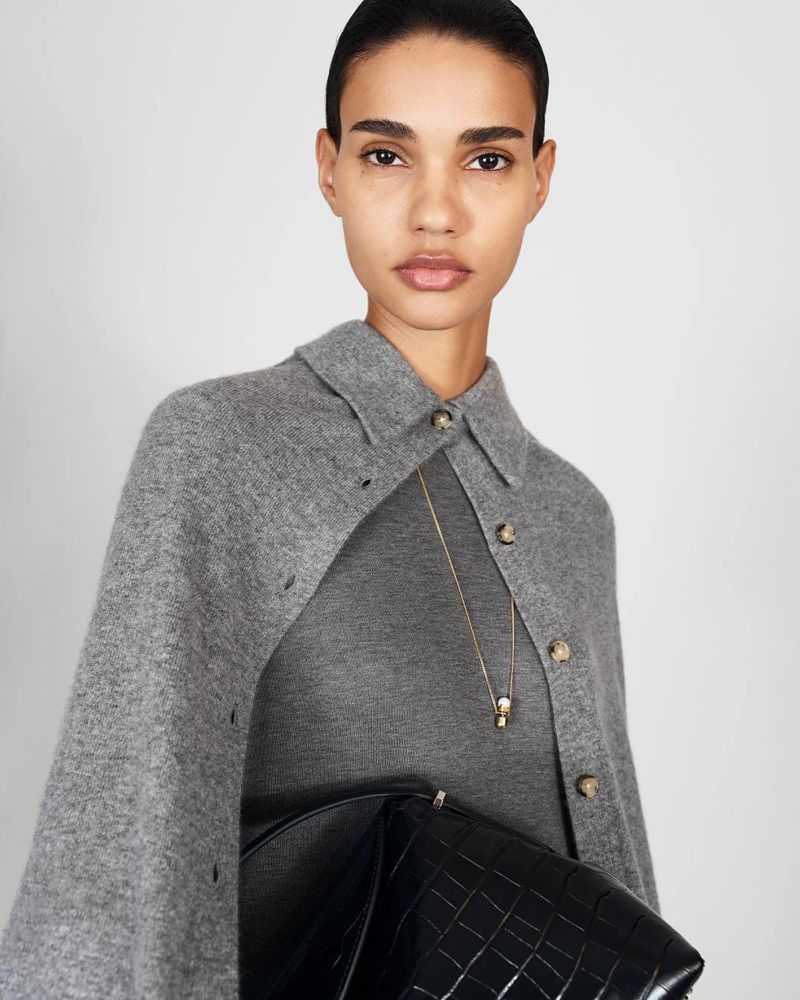 A woman in a grey cape stands indoors holding a black crocodile embossed purse wearing a grey dress and gold necklace against a white background.