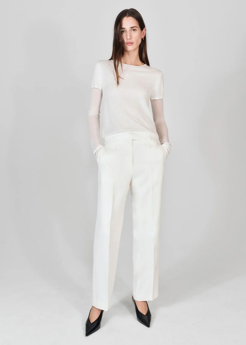Fashionable woman in white top and trousers with black heels posing in studio. Neutral background, full-length shot, studio lighting. 