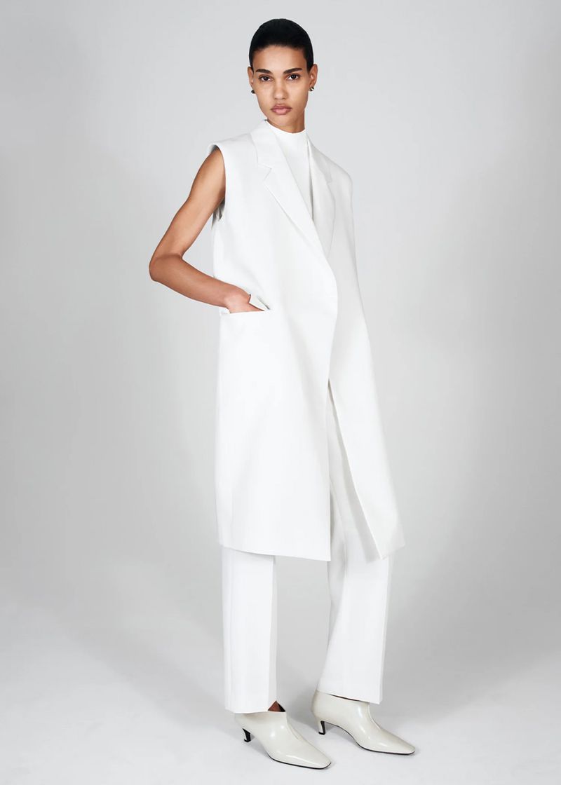Full-length shot of a slender woman in an all-white, minimalist fashion ensemble posing against a neutral backdrop in a studio setting.