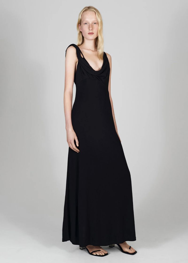Full-length shot of a fair-skinned woman with blonde hair wearing a black, floor-length dress and black sandals against a neutral background.