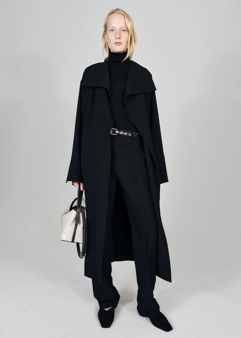 Full-length shot of a slender adult woman in a black coat and pants, holding a cream-and-black purse against a pale grey studio backdrop.