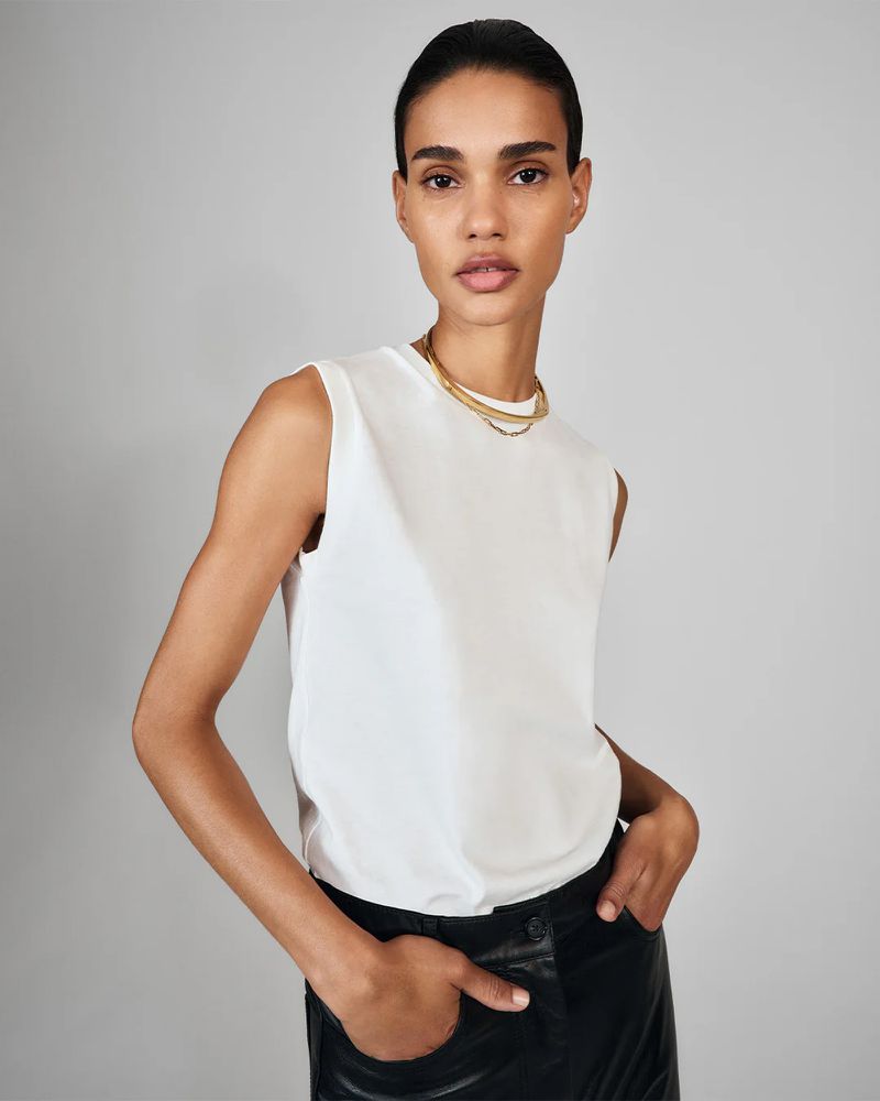 Woman in white top and black pants poses in a studio. Minimalist fashion shot for e-commerce, showing neutral background and simple gold jewelry.