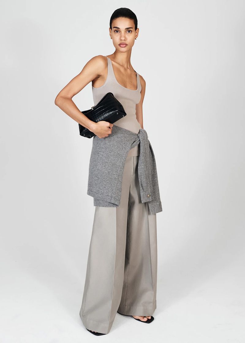 A slender woman poses in a beige bodysuit and wide-leg pants, holding a black clutch and a gray cardigan against a plain background.