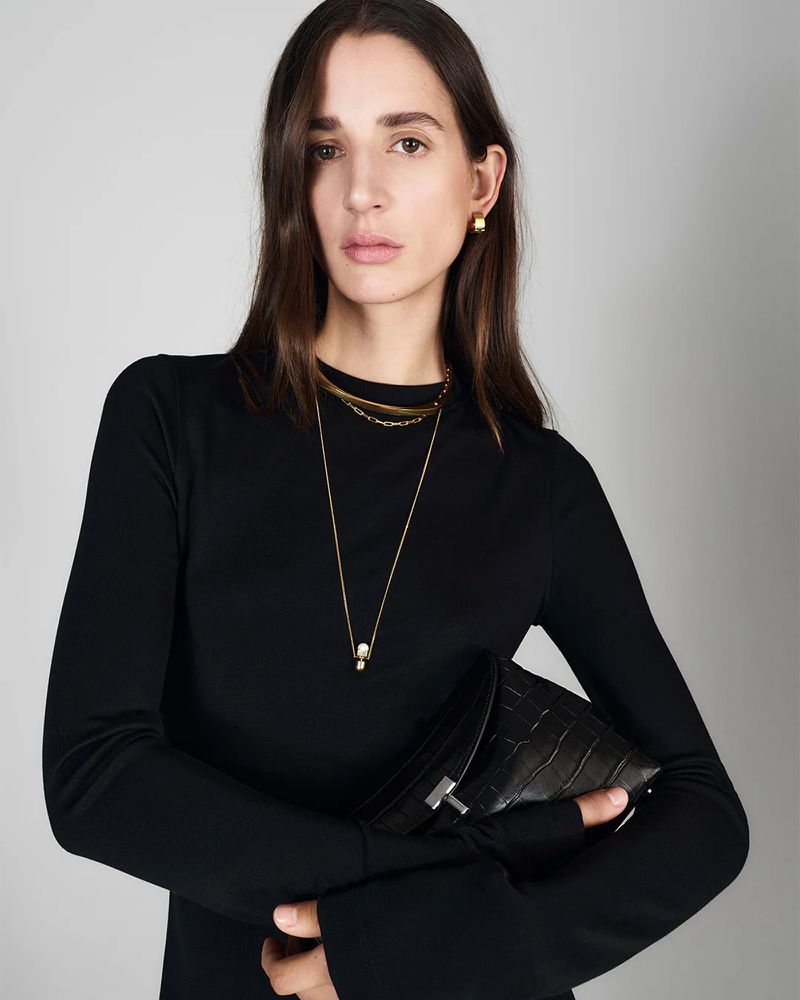 Woman in a black long-sleeved top holds a black alligator-skin clutch. Gold necklace and earrings. Grey background. Fashion photography.