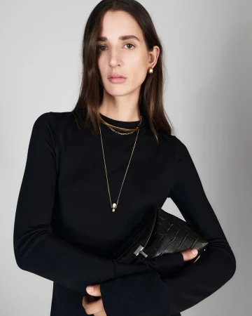 Woman in a black long-sleeved top holds a black alligator-skin clutch. Gold necklace and earrings. Grey background. Fashion photography.
