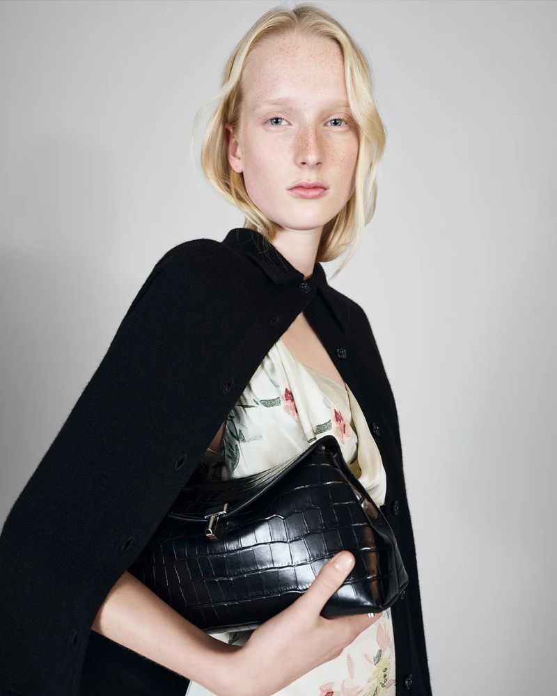 Woman with blonde hair poses indoors wearing a black cape and floral dress, holding a black crocodile-texture leather handbag.