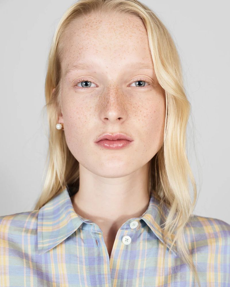 Close-up portrait of a young adult woman with blonde hair and freckles wearing a button-up shirt with a blue and yellow plaid pattern.