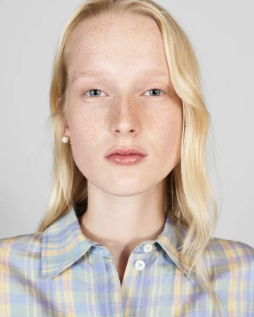 Close-up portrait of a young adult woman with blonde hair and freckles wearing a button-up shirt with a blue and yellow plaid pattern.