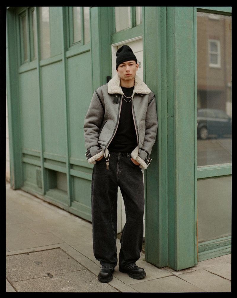 Young man poses outdoors in a gray aviator jacket with black jeans and beanie, leaning against an urban green building in daylight.