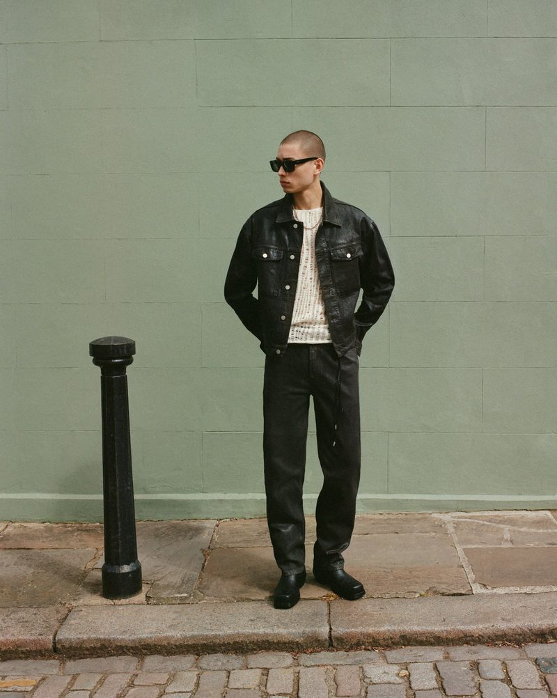 A fashionable adult man in a black denim jacket and matching pants stands in front of a green wall on a cobblestone street, sunglasses.