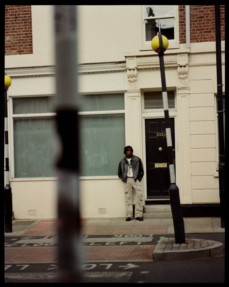 An adult male poses on a city sidewalk in front of a building with a dark door and a crosswalk in an overcast urban setting.