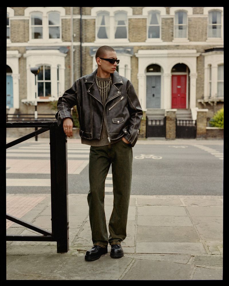A man in a leather jacket stands on a city street near brick buildings, a crosswalk, and a black metal railing on a cloudy day.