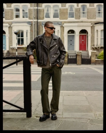 A man in a leather jacket stands on a city street near brick buildings, a crosswalk, and a black metal railing on a cloudy day.