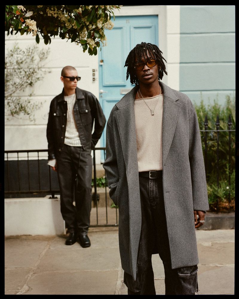 Two stylish adults strike poses on a city street, one in a grey coat and the other in a denim jacket with sunglasses, exuding urban fashion.